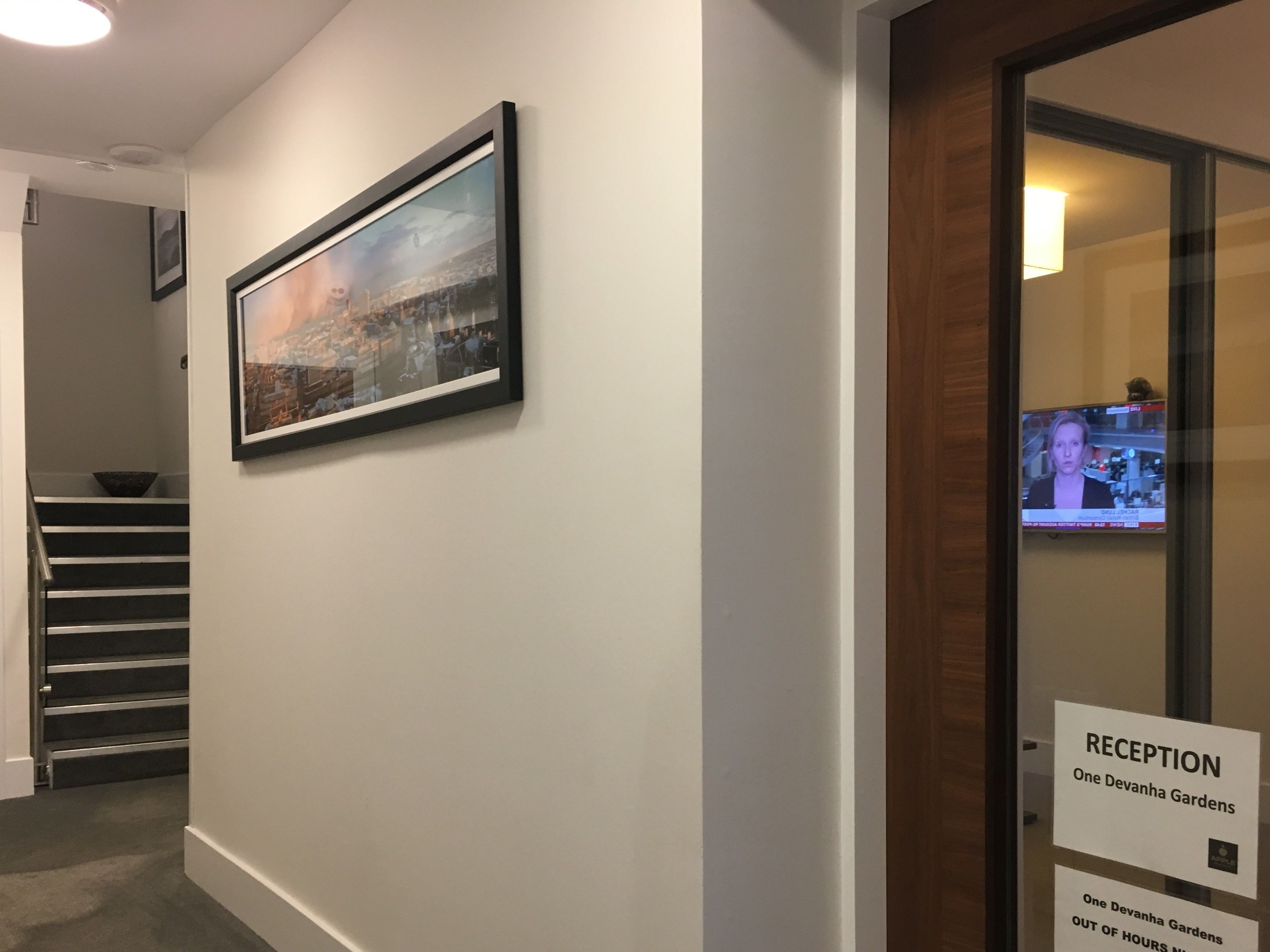 A reception area with a framed cityscape painting, a staircase, and a television displaying a news channel.
