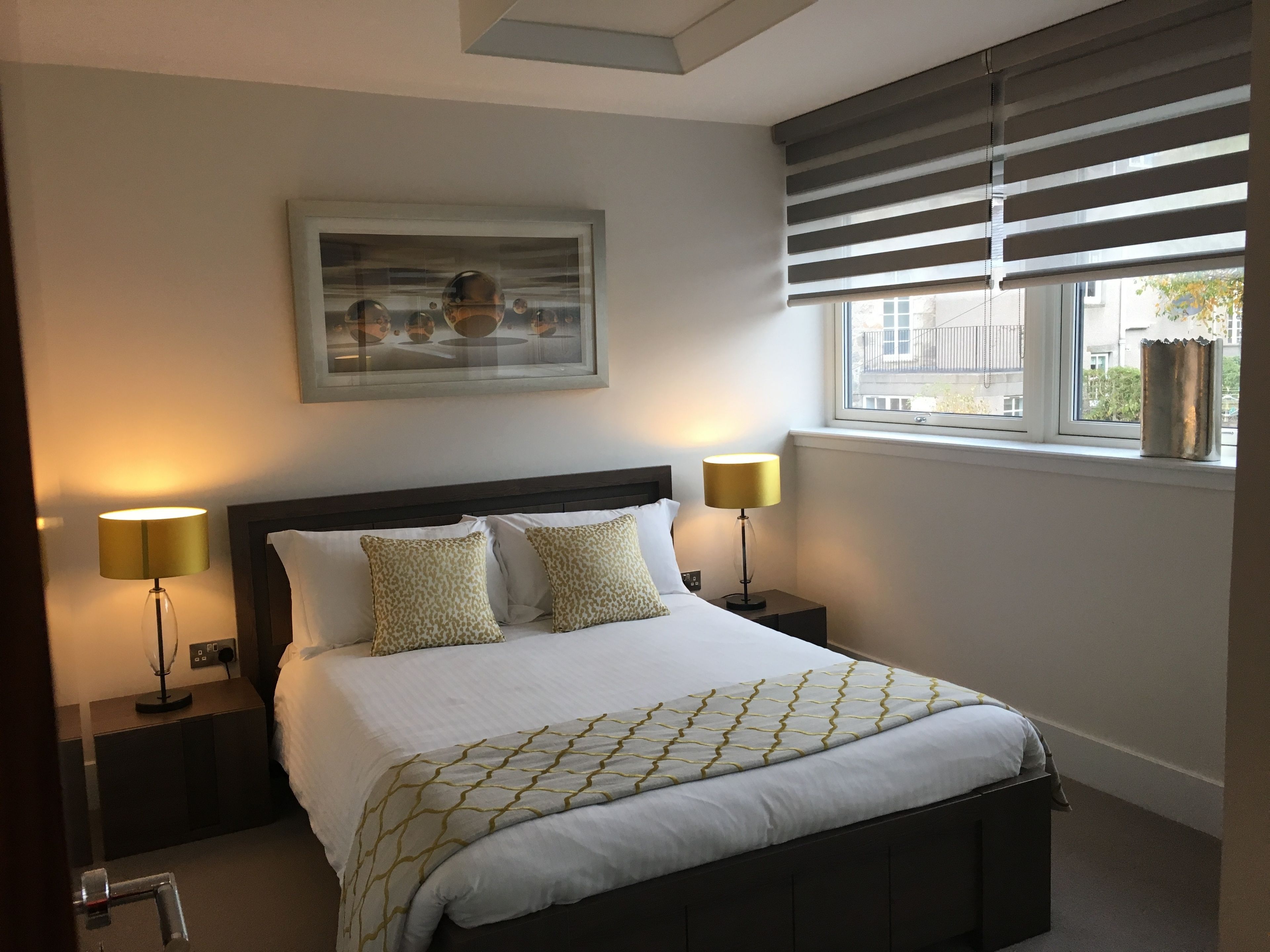 A neatly arranged bedroom with a bed, two bedside lamps, a window with blinds, and a framed picture on the wall.