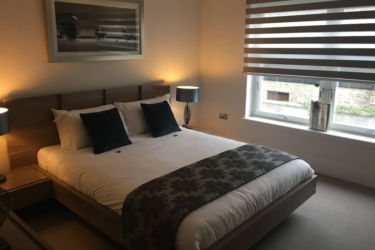 Une chambre à coucher avec un grand lit, deux lampes de chevet, une fenêtre avec des stores et un tableau encadré au mur.