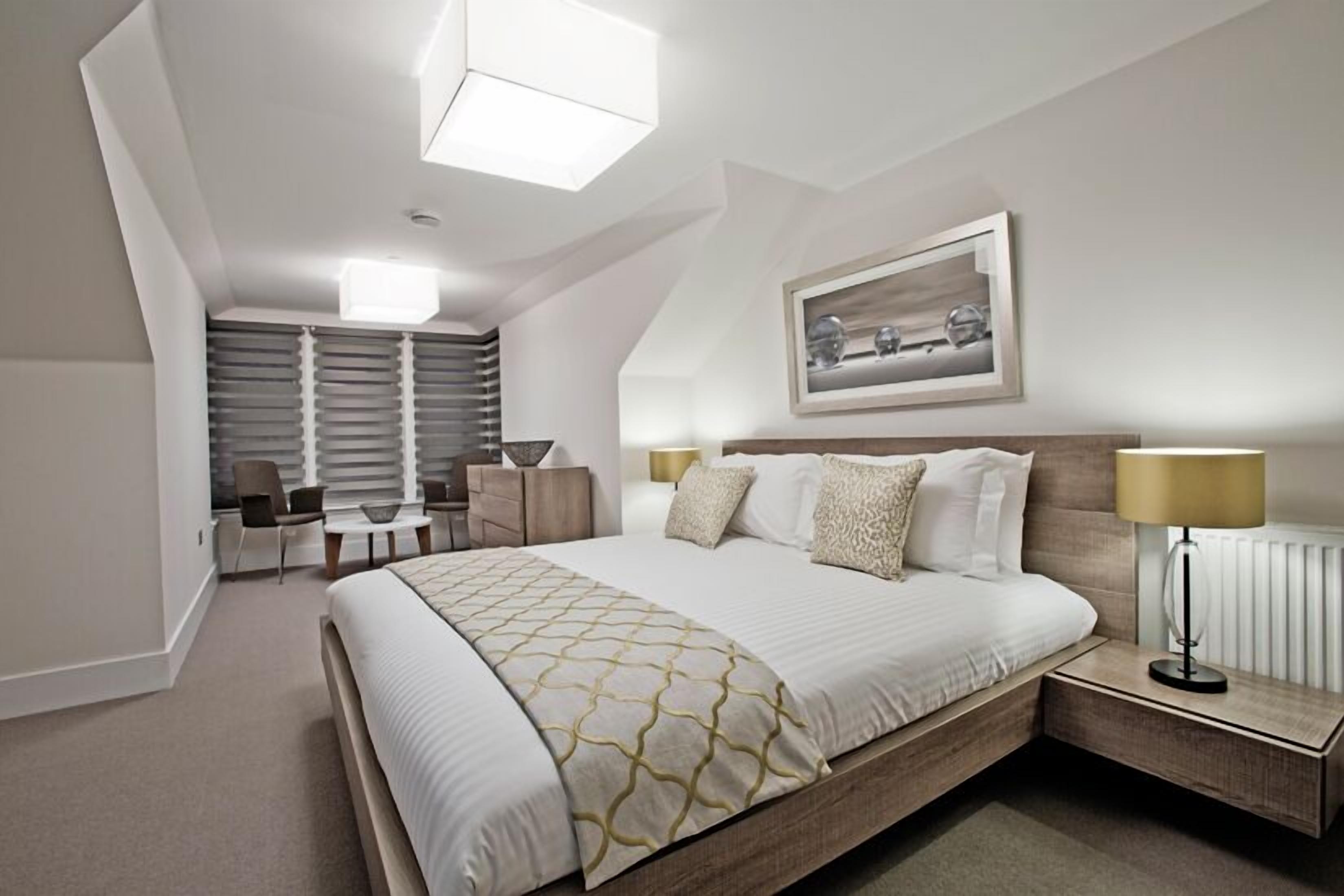 A modern hotel room with a large bed, a desk, a chair, and a bedside table with a lamp.