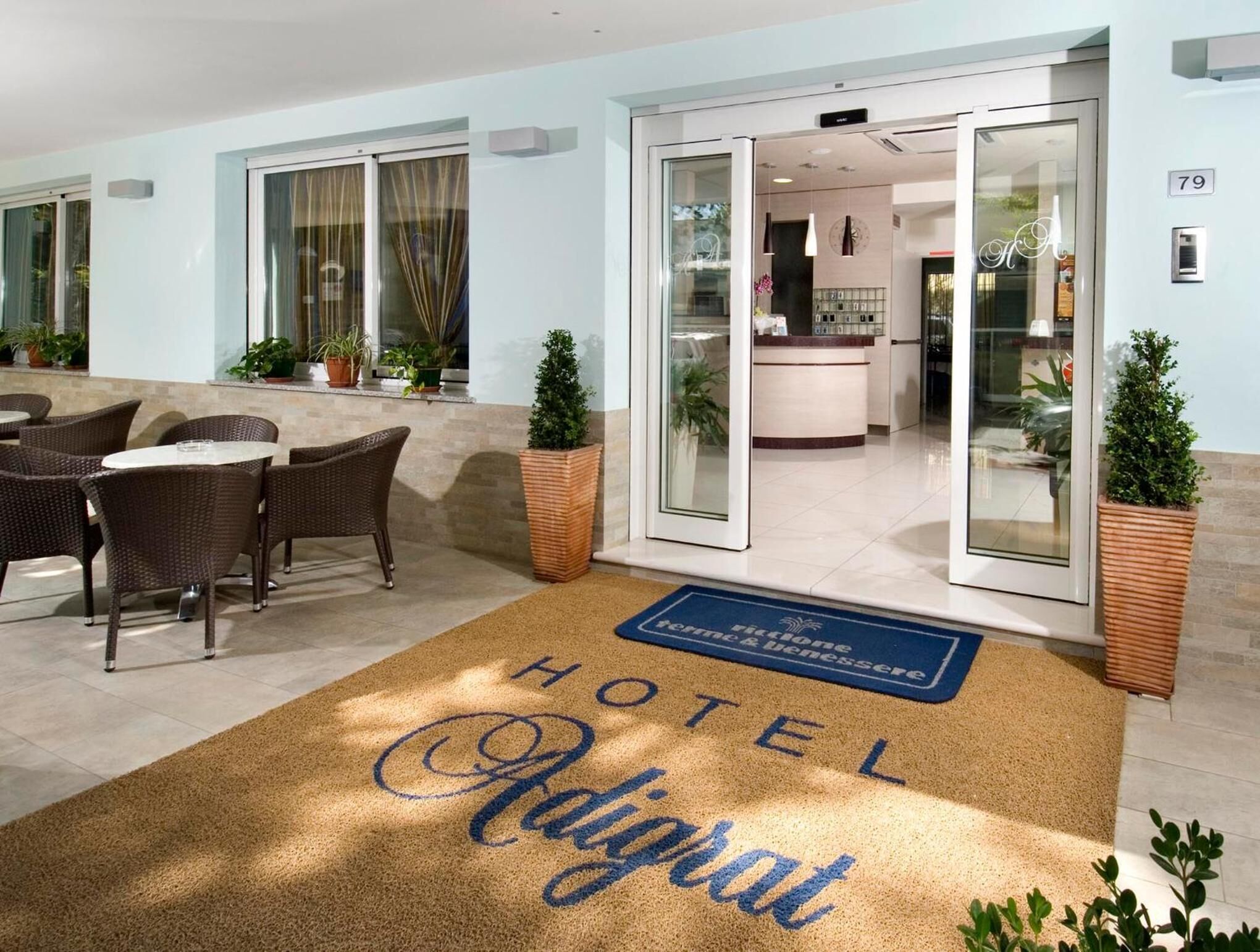 L'ingresso di un hotel con una reception, un'area salotto con mobili in vimini e un tappetino con il nome dell'hotel.