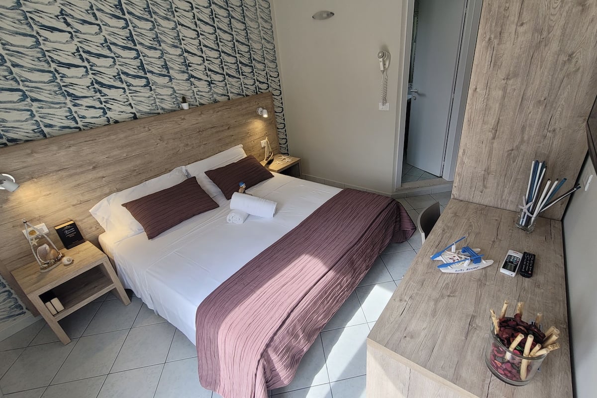 A hotel room with a wooden headboard, a bed with white and brown bedding, a nightstand with a lamp, a small table with a remote control, a glass of wine, and a bottle of water.