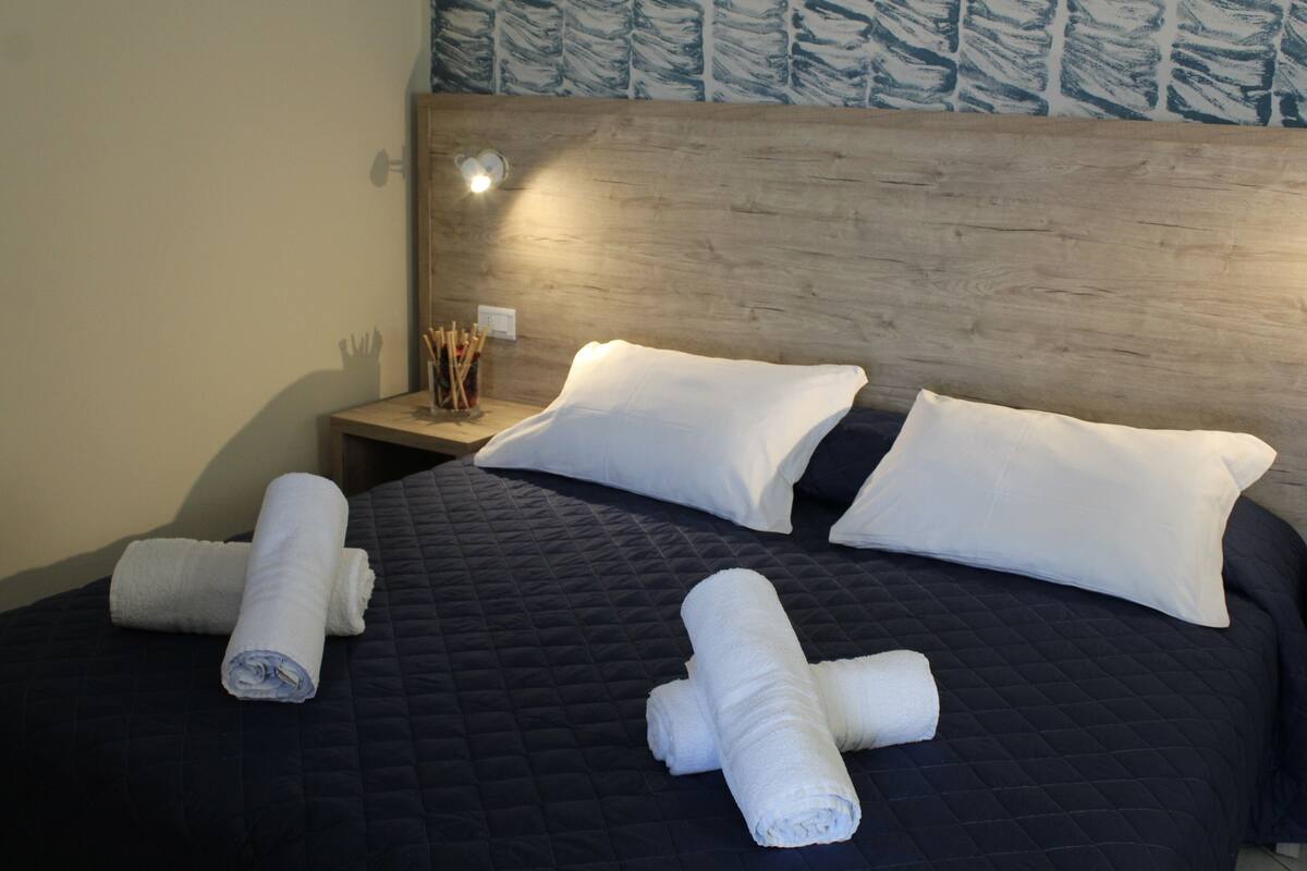 A neatly made bed with a dark quilt, two white pillows, and rolled towels on the bed.