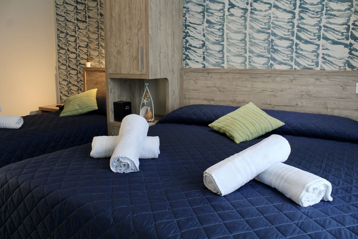 A hotel room with two beds, each with a dark blue quilted bedspread and white towels rolled up. There is a wooden headboard, a small wooden shelf with a lamp, and a wall with a leaf pattern.