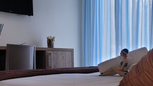 A bedroom with a bed, a wooden nightstand, a television, and a window with blue curtains. - Hotel Adigrat (Riccione)