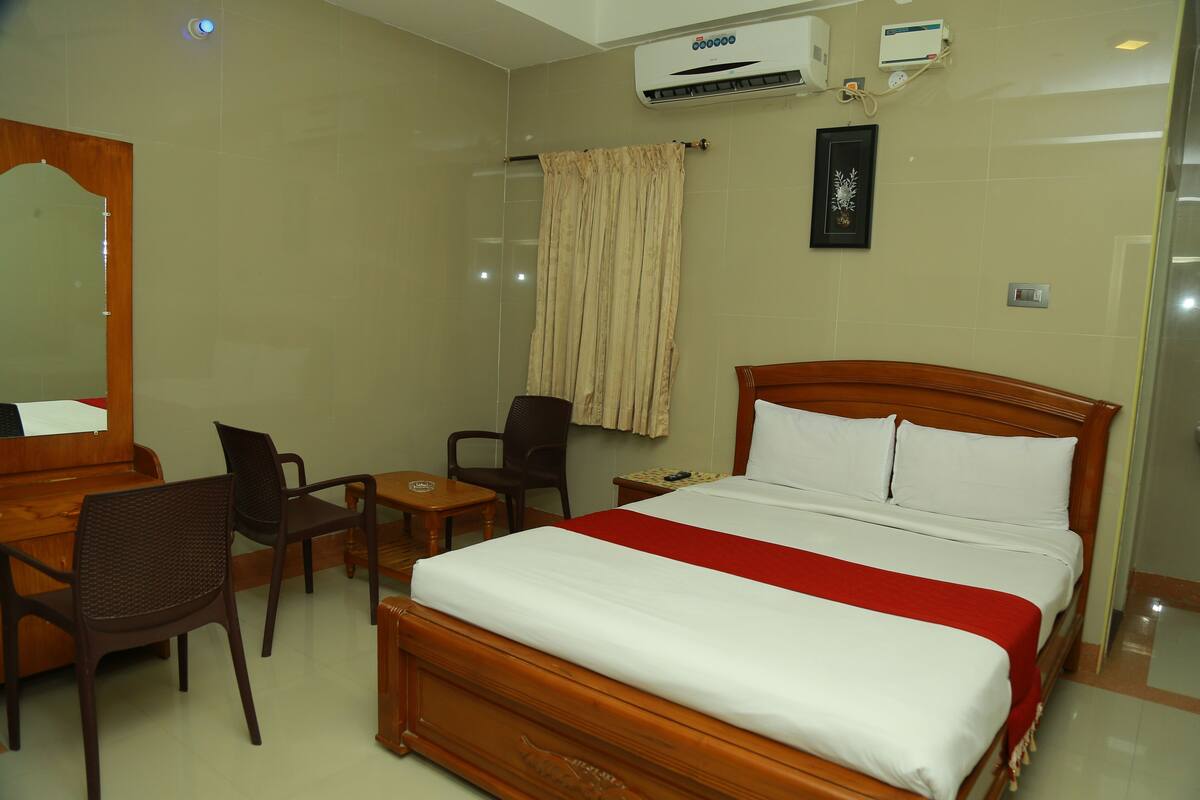 A hotel room with a wooden bed, a desk, two chairs, a small table, and a wall-mounted air conditioner.
