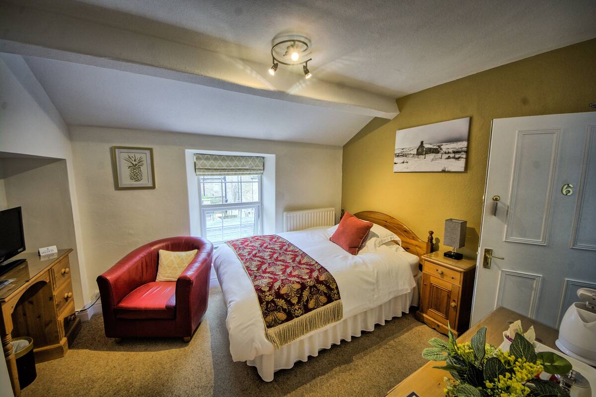 A bedroom with a bed, a red armchair, a wooden nightstand, a television, and a window with blinds.