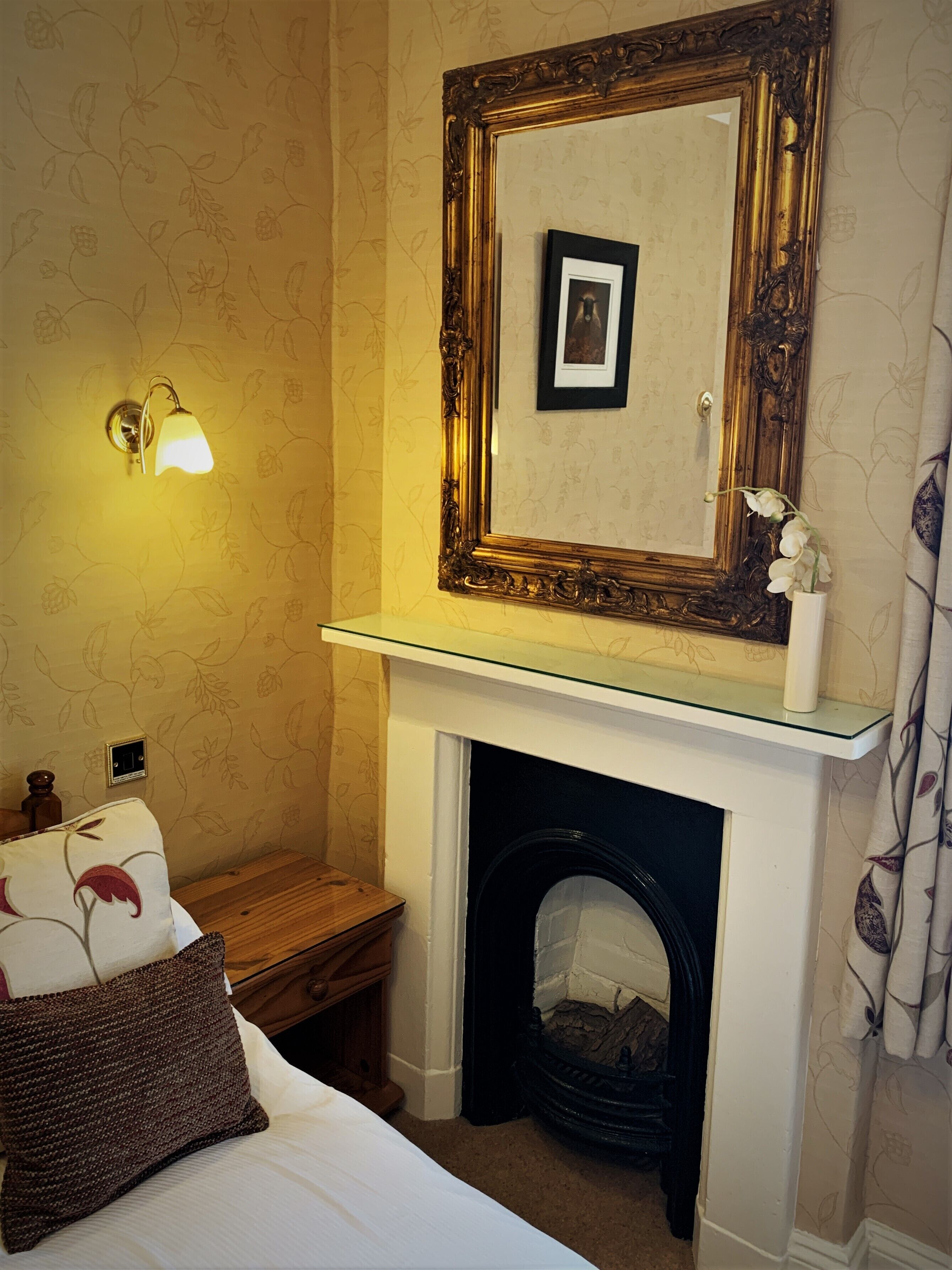 Una camera d'albergo con camino, uno specchio con cornice decorata, un comodino con lampada e una parete con motivo floreale.