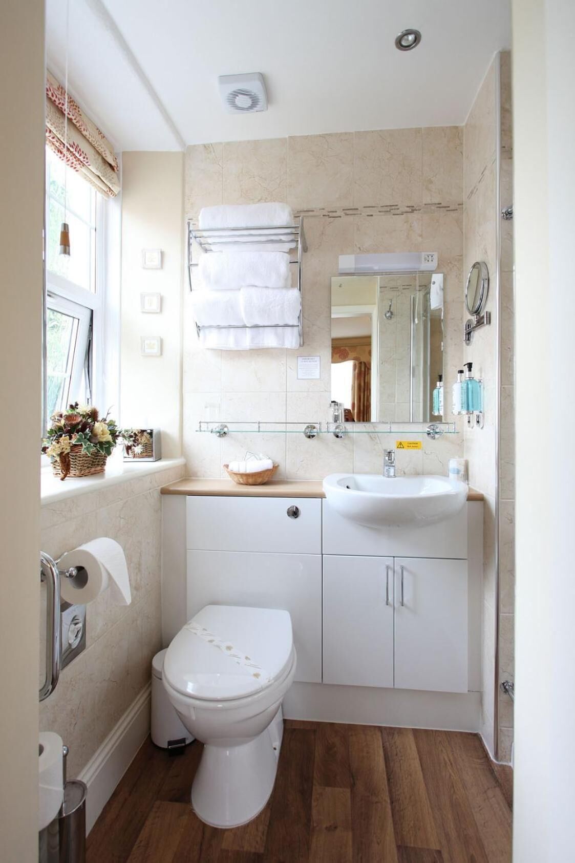 Un baño moderno con inodoro, lavamanos y espejo. Hay una ventana con cortinas, un perchero para toallas y una cesta con flores.