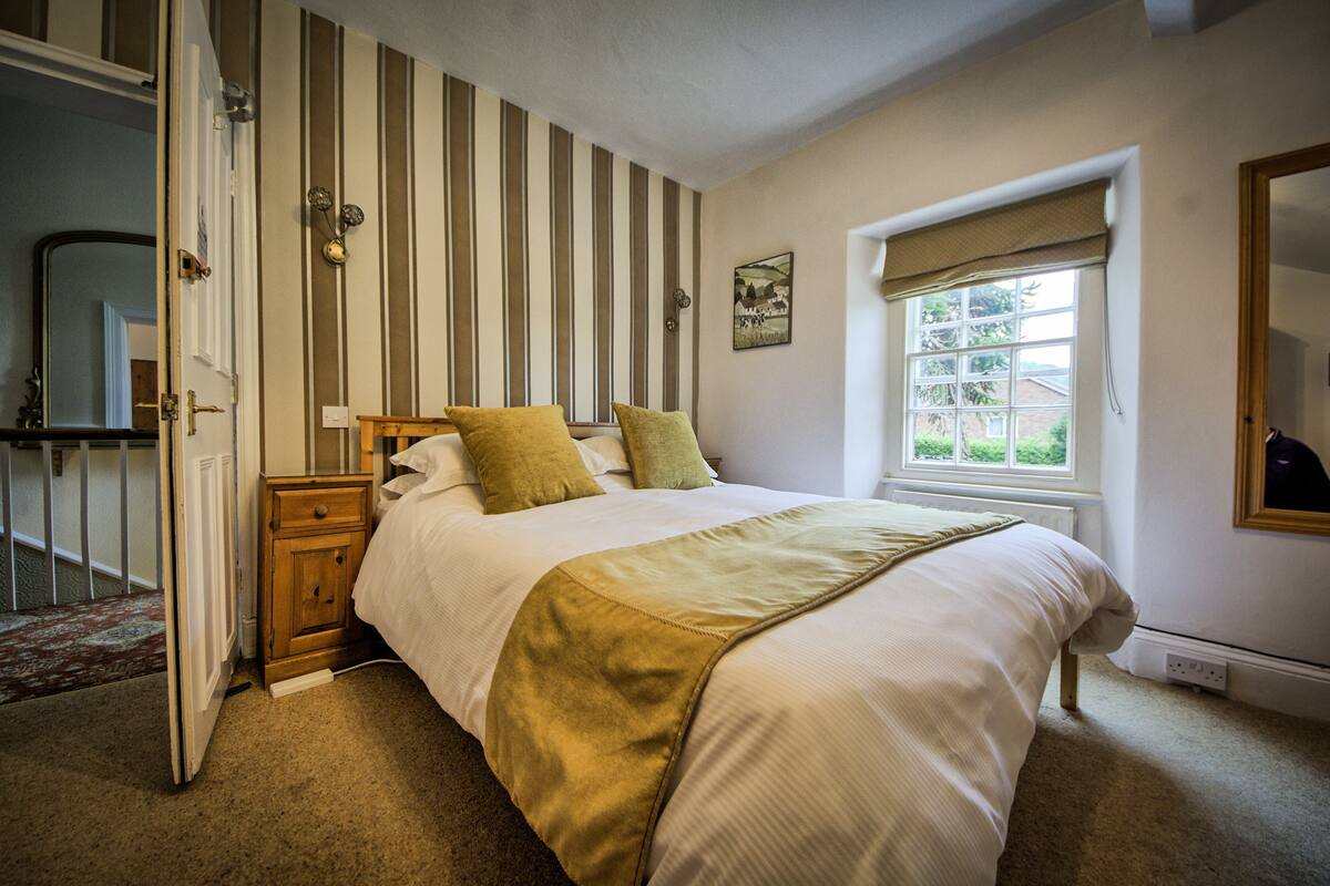 A bedroom with a bed, a nightstand, a window with a view, and a door leading to another room.