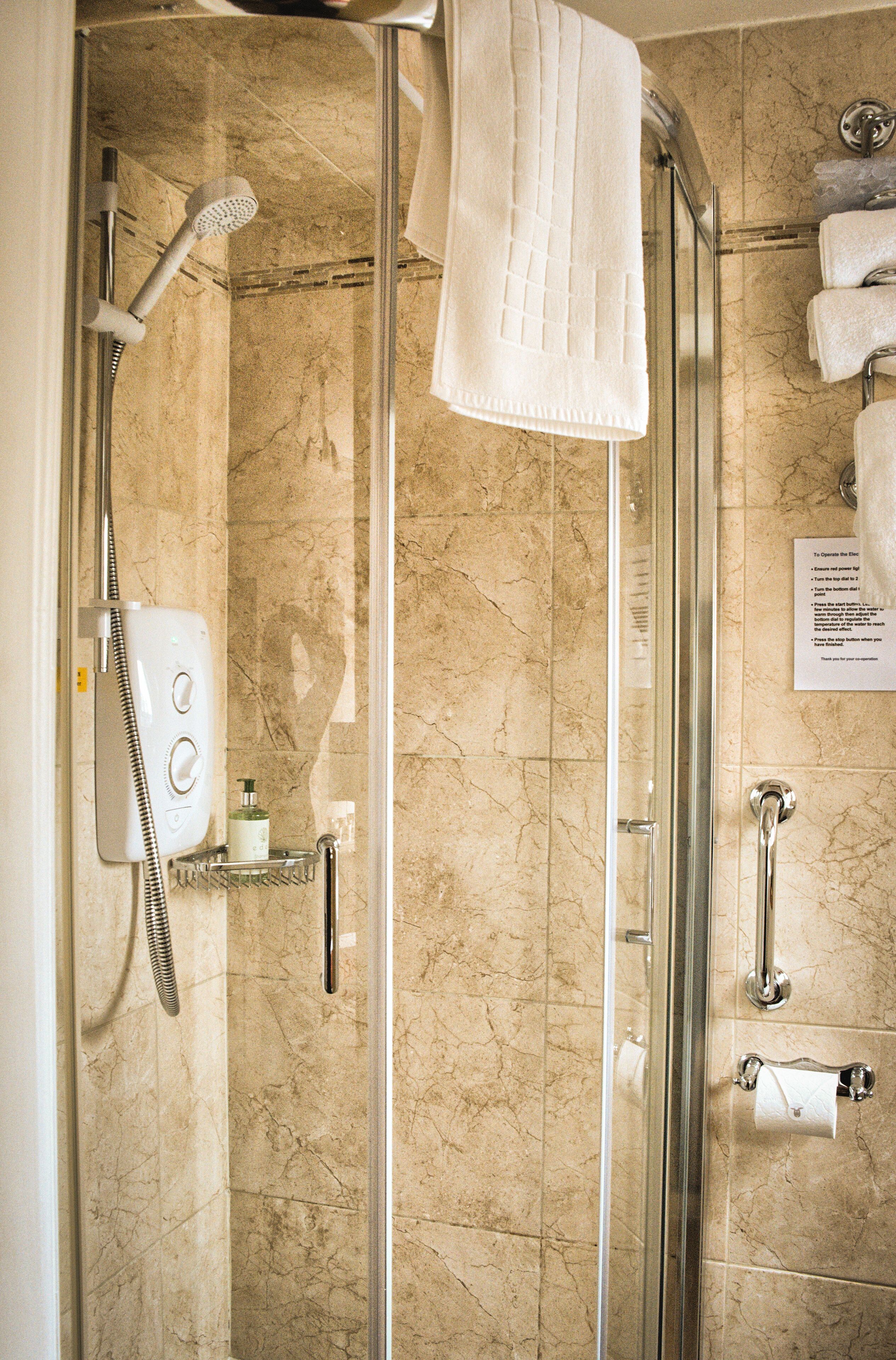 Une douche fermée par des parois en verre, dotée d’un pommeau de douche à main, d’un porte-serviettes avec une serviette pliée, d’un distributeur de savon et d’une petite étagère contenant des articles de toilette.