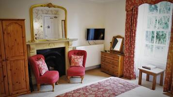 Une pièce avec une cheminée, deux fauteuils rouges, un meuble en bois, une commode, un téléviseur et une fenêtre avec des rideaux.