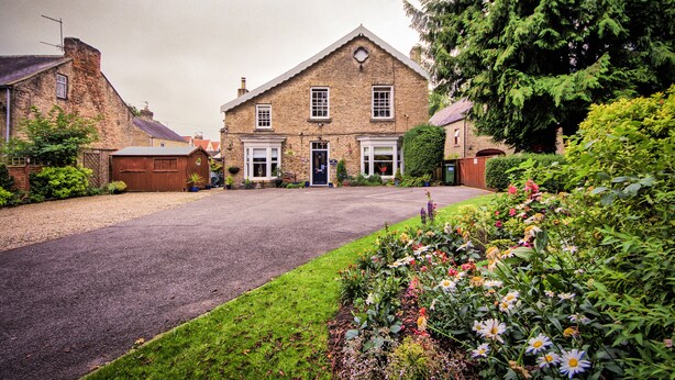 A two-story stone house with a front driveway, a wooden garage door, and a well-maintained garden with various flowers. - West End Guest House (Richmond)