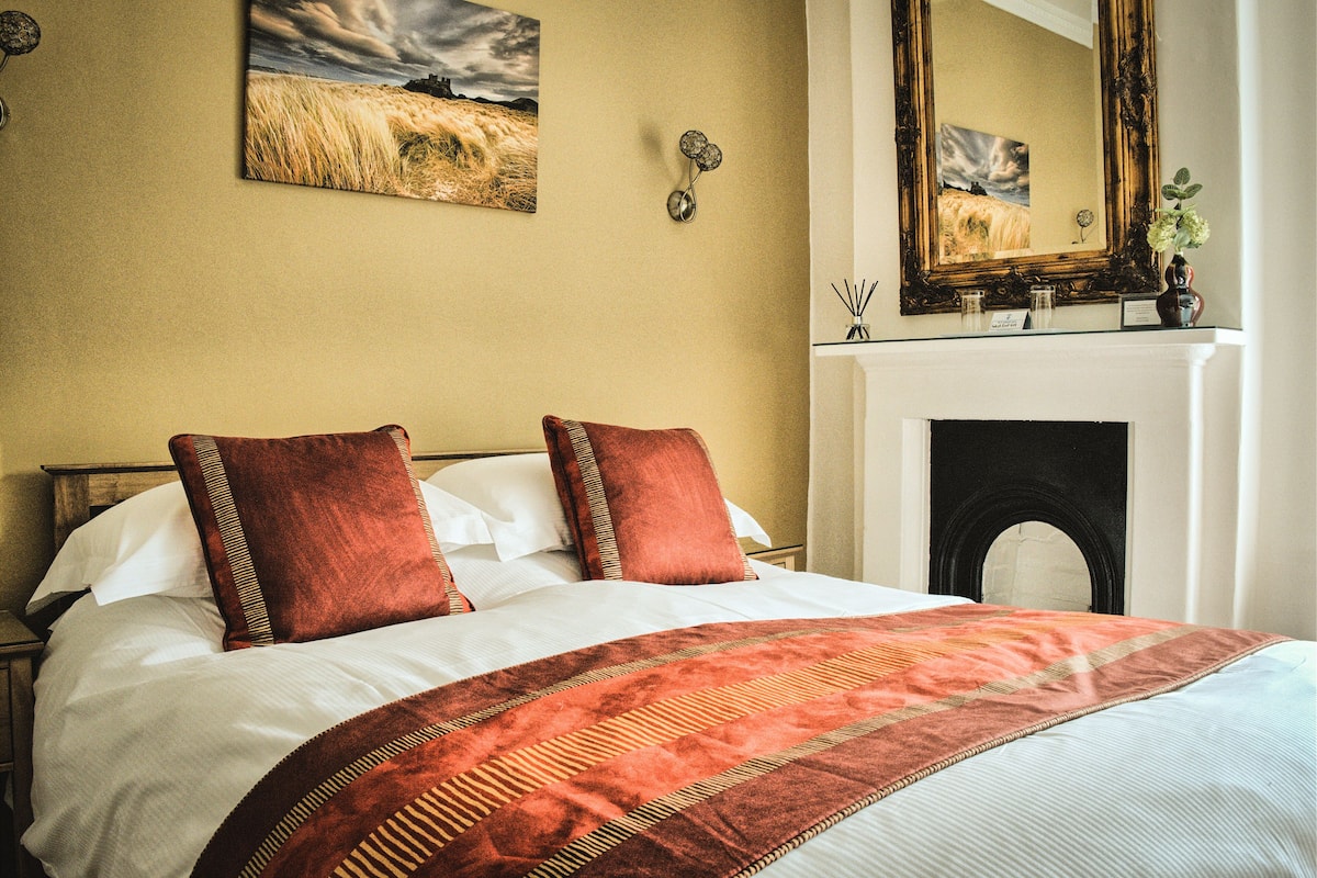 A neatly made bed with a patterned bedspread, a large mirror above the fireplace, and a painting on the wall.