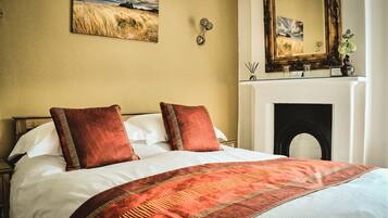 A neatly made bed with a patterned bedspread, a large mirror above the fireplace, and a painting on the wall.