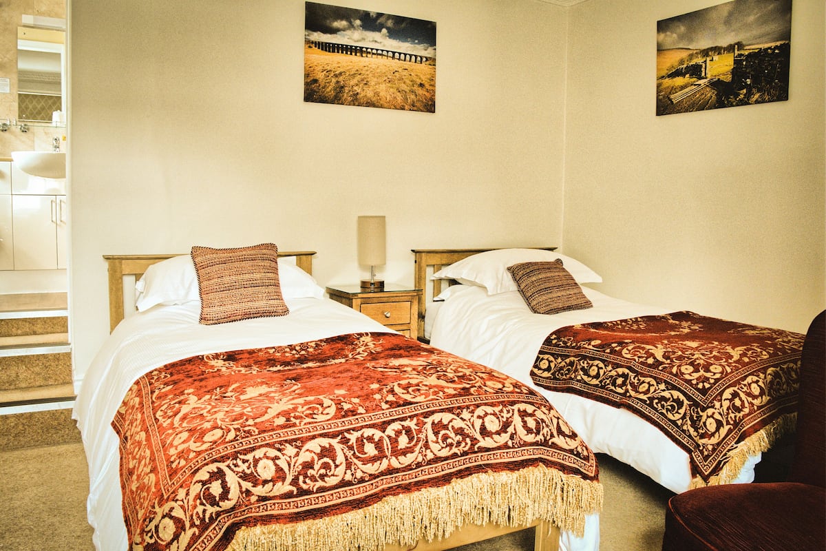 A hotel room with two beds, each with a patterned bedspread and pillows, a nightstand with a lamp, and a wall-mounted picture.
