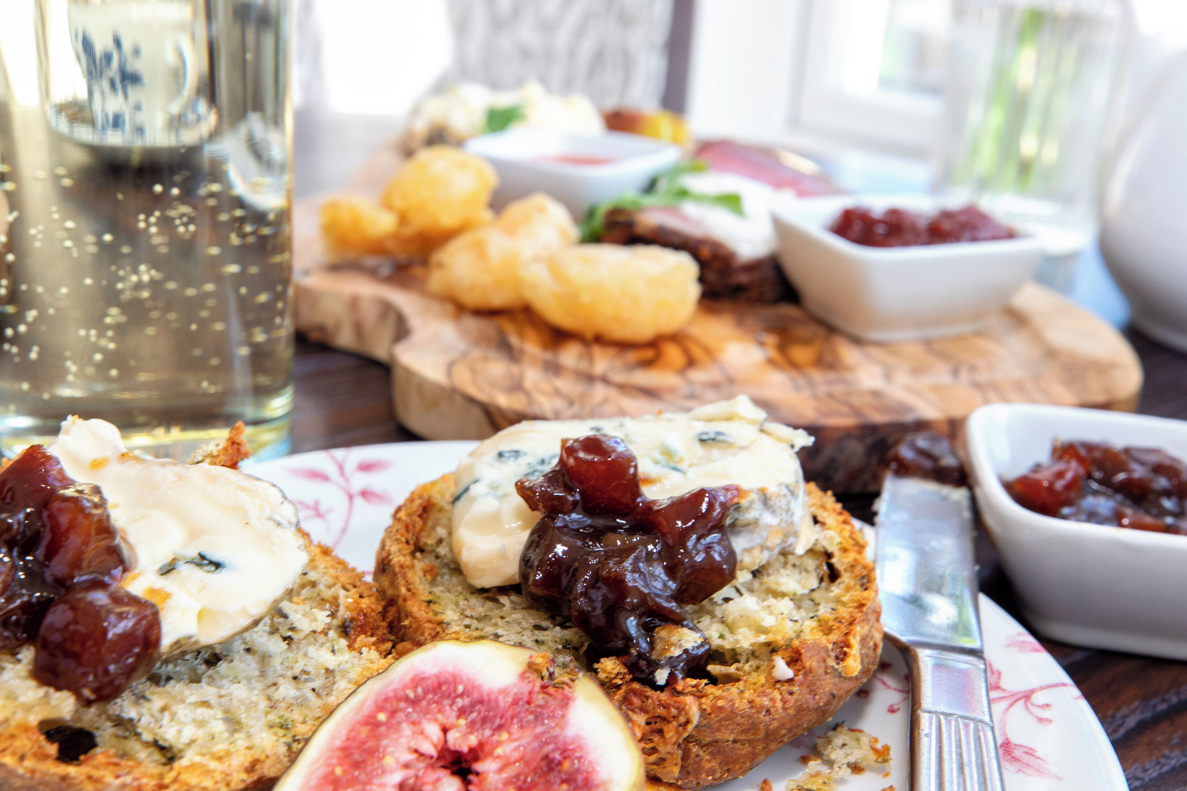 Una mesa con diversos alimentos, incluyendo pan con queso e higos, un bol de mermelada y una botella de bebida.