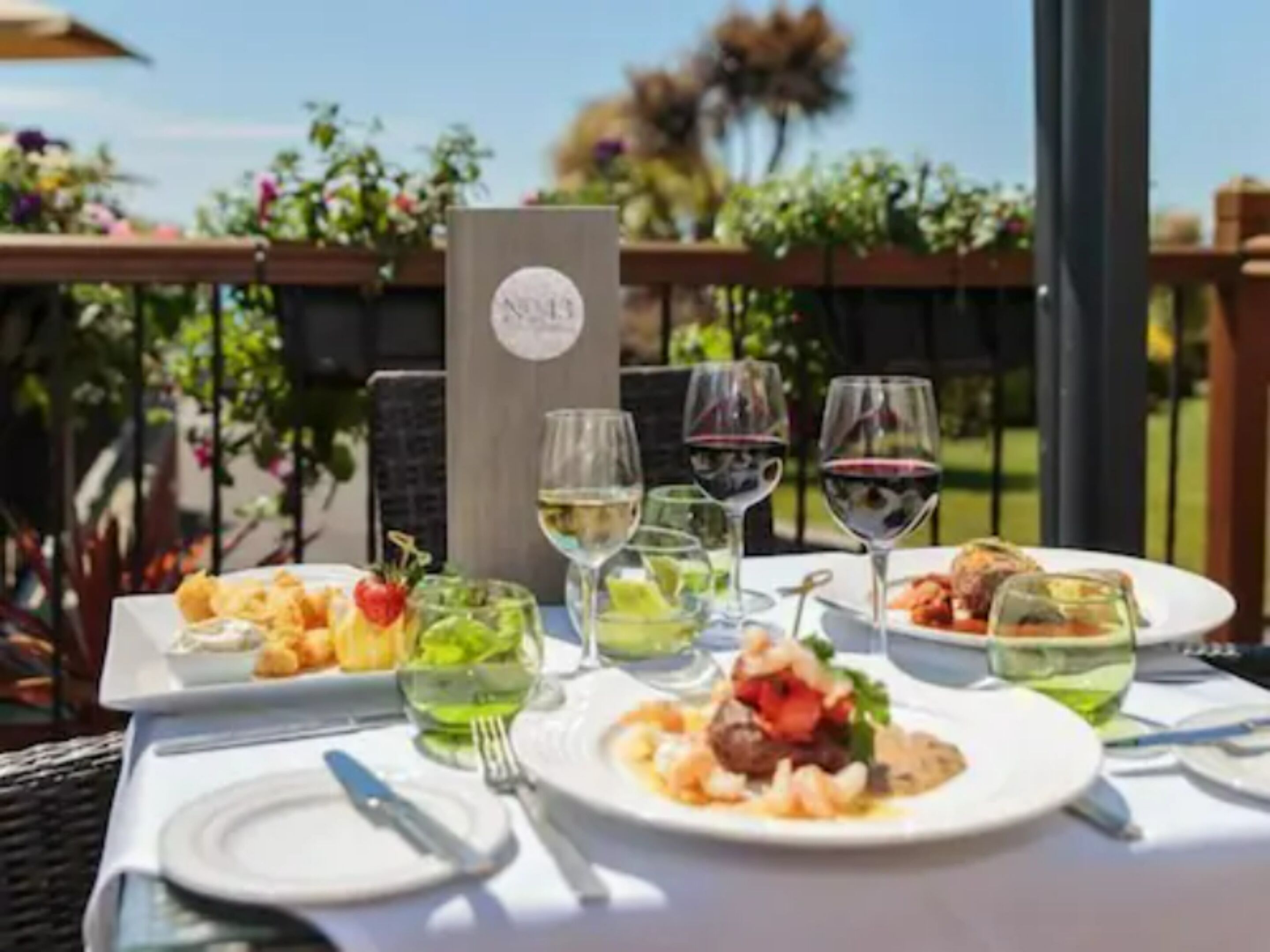 A table set for a meal with various dishes and glasses of wine, outdoors with a view of greenery.