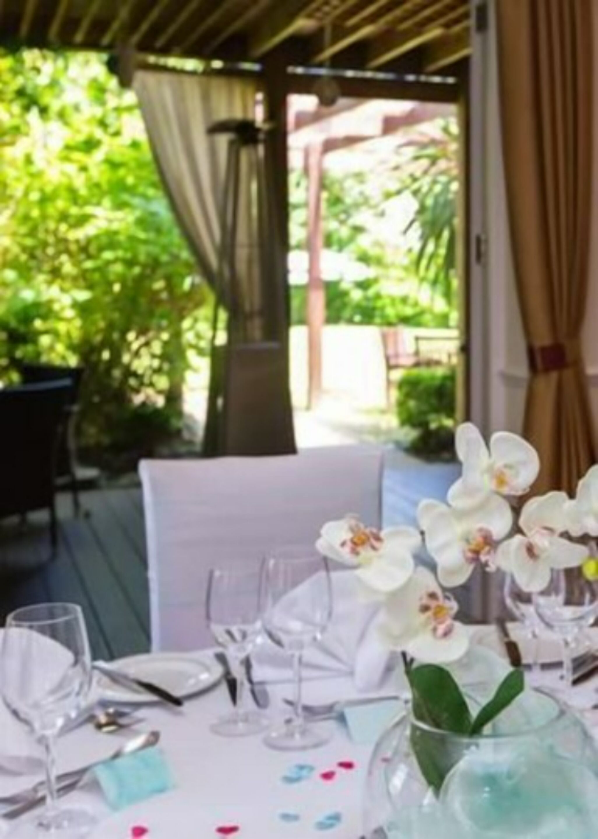Un área de comedor al aire libre con mesas dispuestas para una comida, rodeada de follaje y una pérgola de madera.