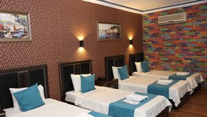 Quarto de hotel com quatro camas individuais, cada uma com uma colcha branca e toalhas azuis. Há duas unidades de ar condicionado fixadas na parede, uma em cada lado do quarto. As paredes são decoradas com papel de parede estampado e papel de parede que imita tijolos coloridos.