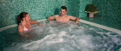 A couple in a hot tub with a tiled wall and a potted plant.