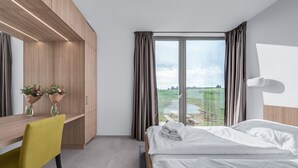 A modern hotel room with a bed, a desk with a chair, a large window with curtains, and a view of a green landscape. - Apartmány Hrádeček (Mlade Buky)