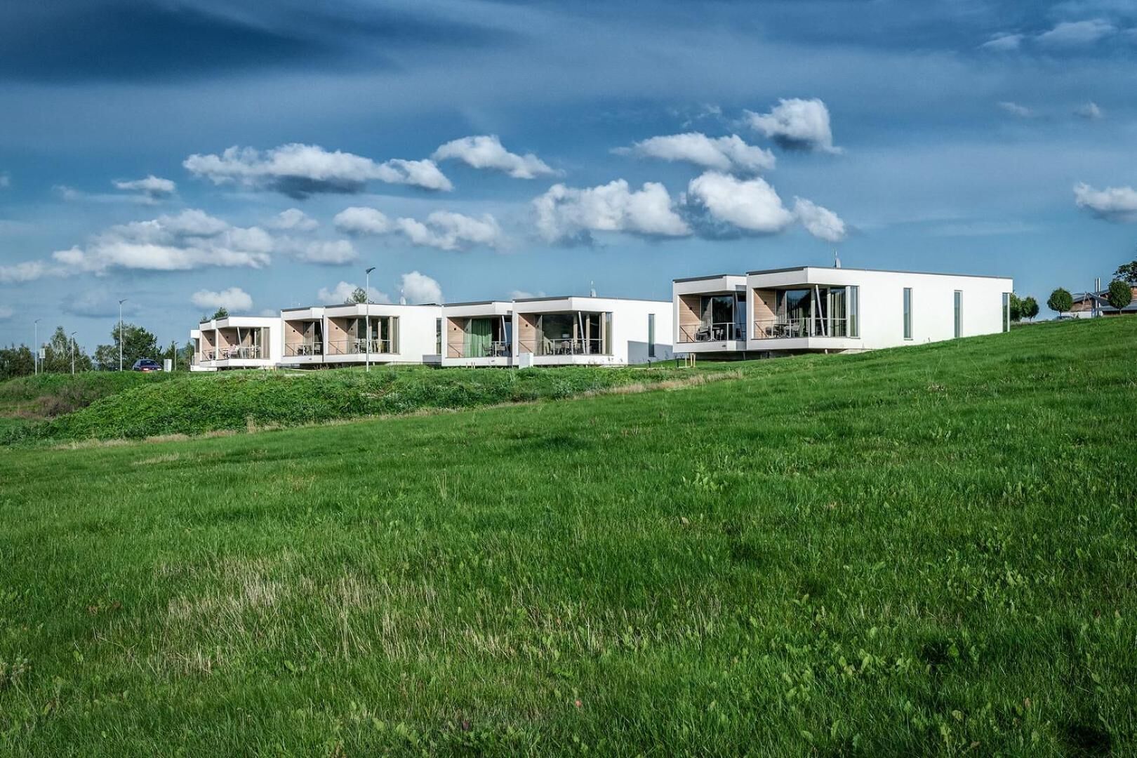 A row of modern, white, two-story houses with large windows and balconies, set on a grassy hill under a clear sky.