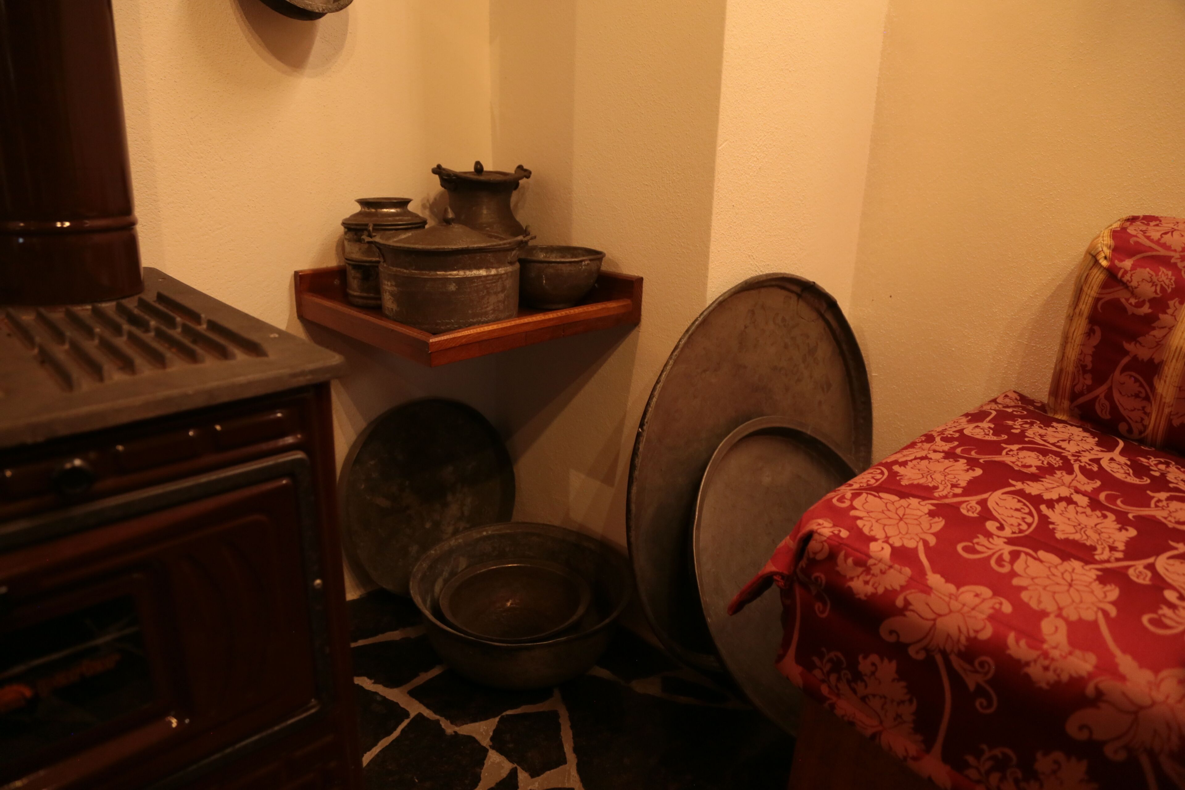 A corner of a room with a wooden cabinet, a shelf with pots, a bowl, and a patterned red bedspread.
