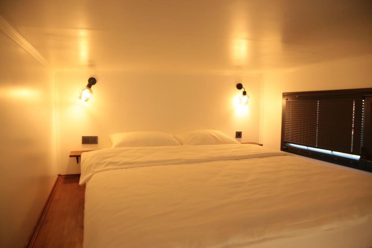 A room with a bed, two wall-mounted light fixtures, a window with blinds, and a wooden floor.