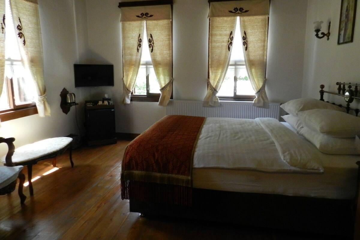 A bedroom with a bed, two windows with curtains, a bedside table, a chair, and a wall-mounted TV.