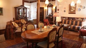 Un comedor con una mesa de madera, sillas y un mantel de encaje. Hay un sofá con una manta estampada y una araña colgante.