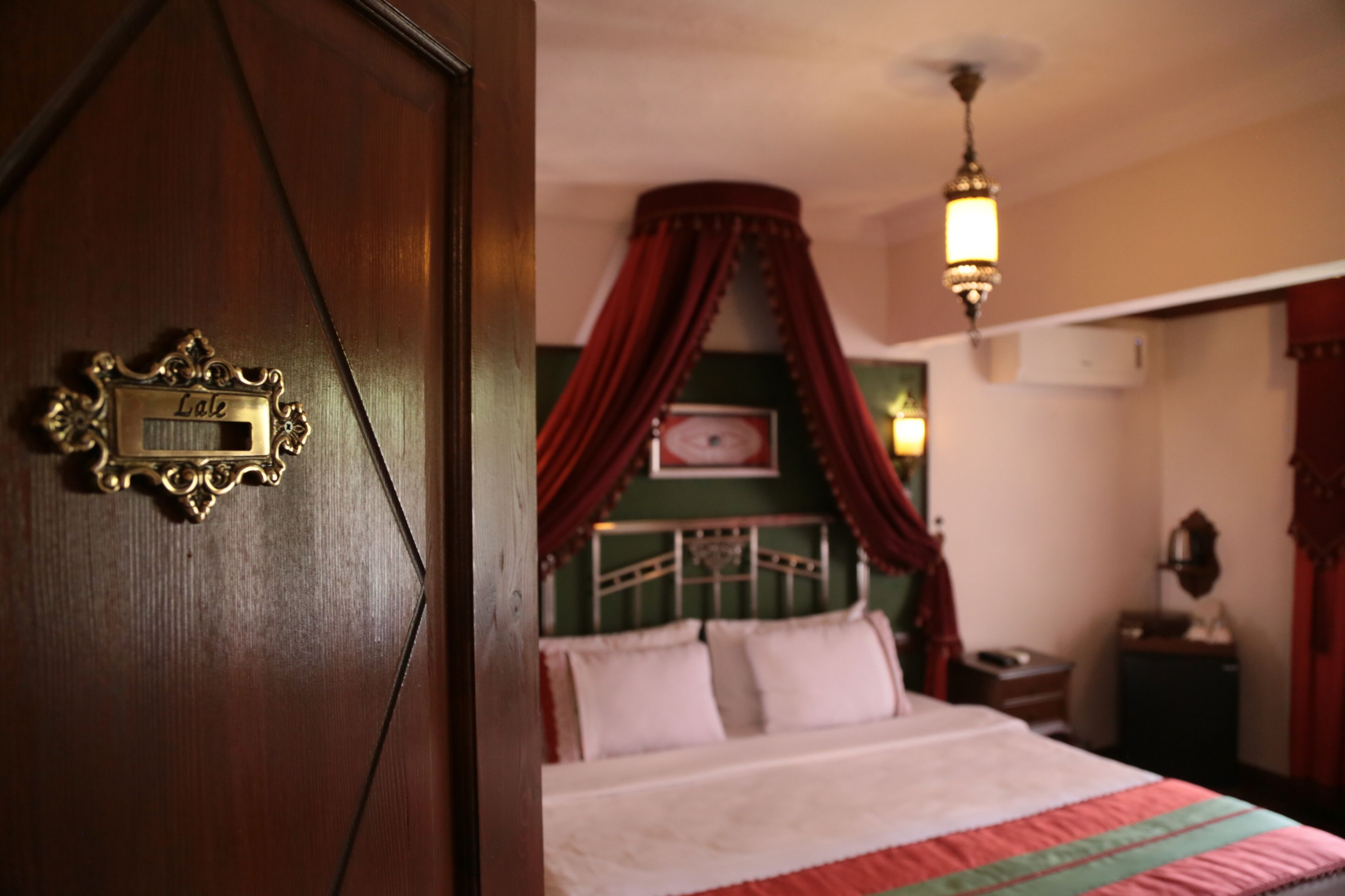 Un dormitorio con cama, una mesita de noche, una lámpara y una puerta de madera con placa.