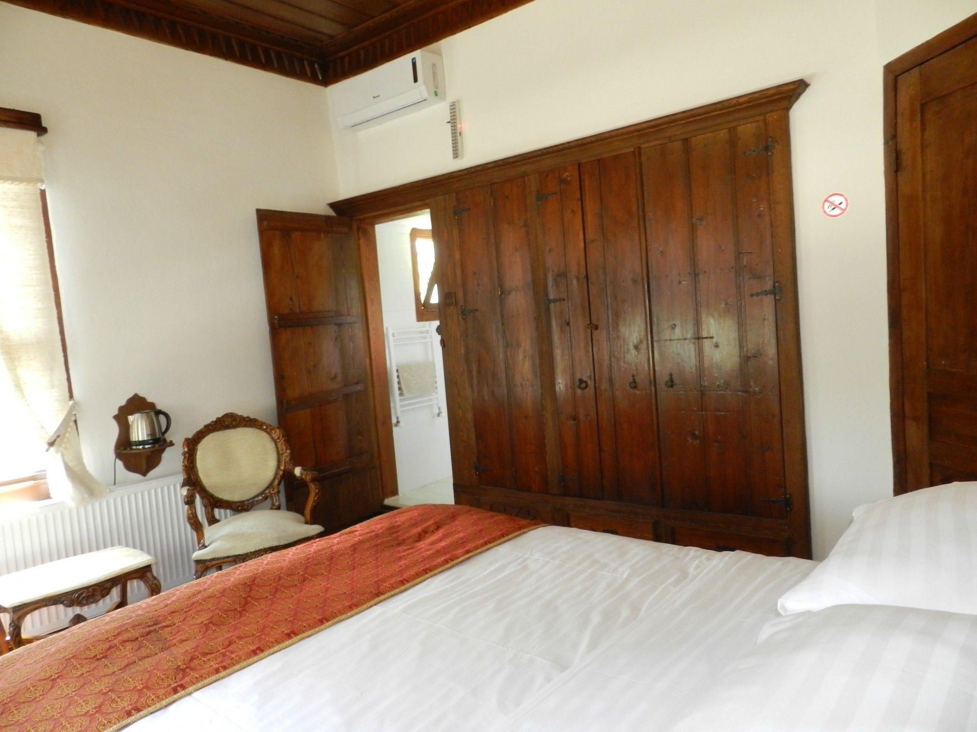 Une chambre avec une grande armoire en bois, un lit avec du linge de lit blanc, une chaise en bois et un radiateur.