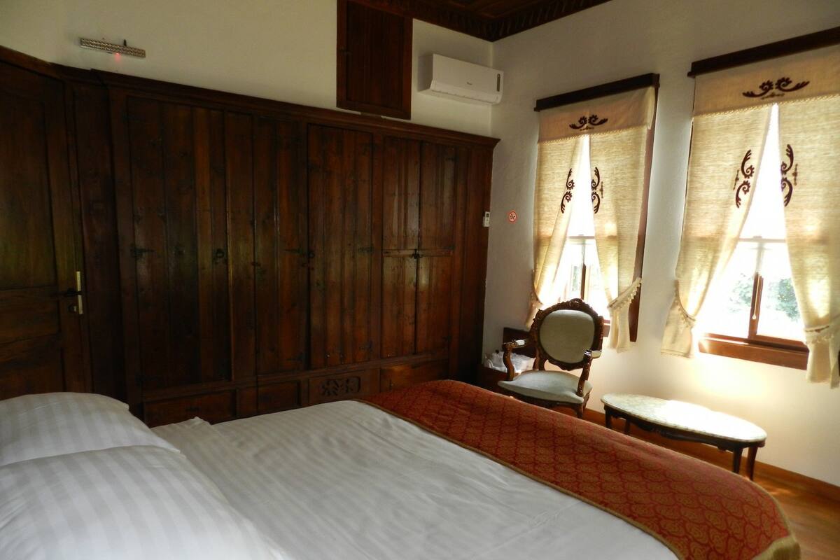 A bedroom with a large wooden wardrobe, a bed with white bedding, a chair, and two windows with curtains.
