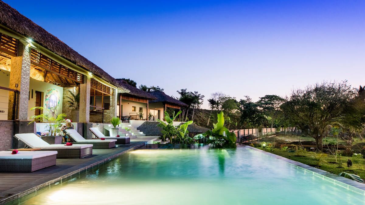 A poolside area with lounge chairs, a thatched-roof building, and lush greenery.