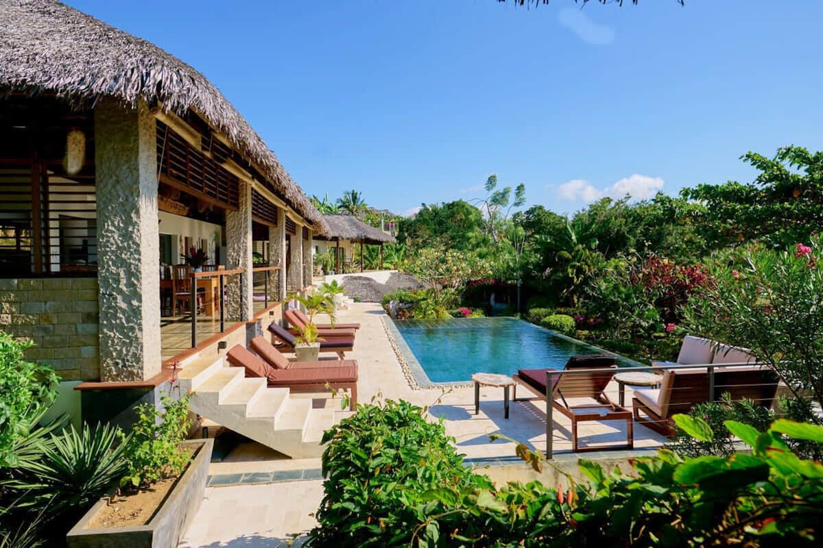 A poolside area with lounge chairs, a thatched-roof building, and lush greenery.