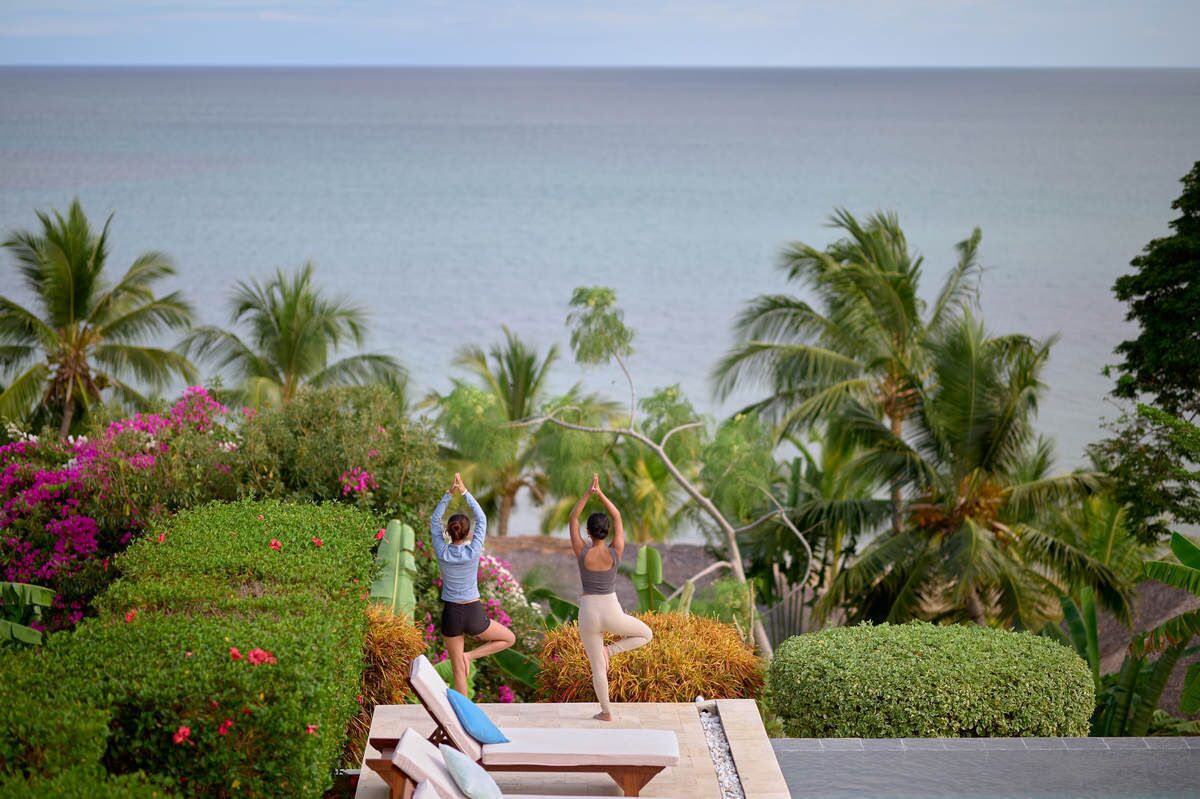 Twee personen doen yoga op een terras met uitzicht op de oceaan en palmbomen.