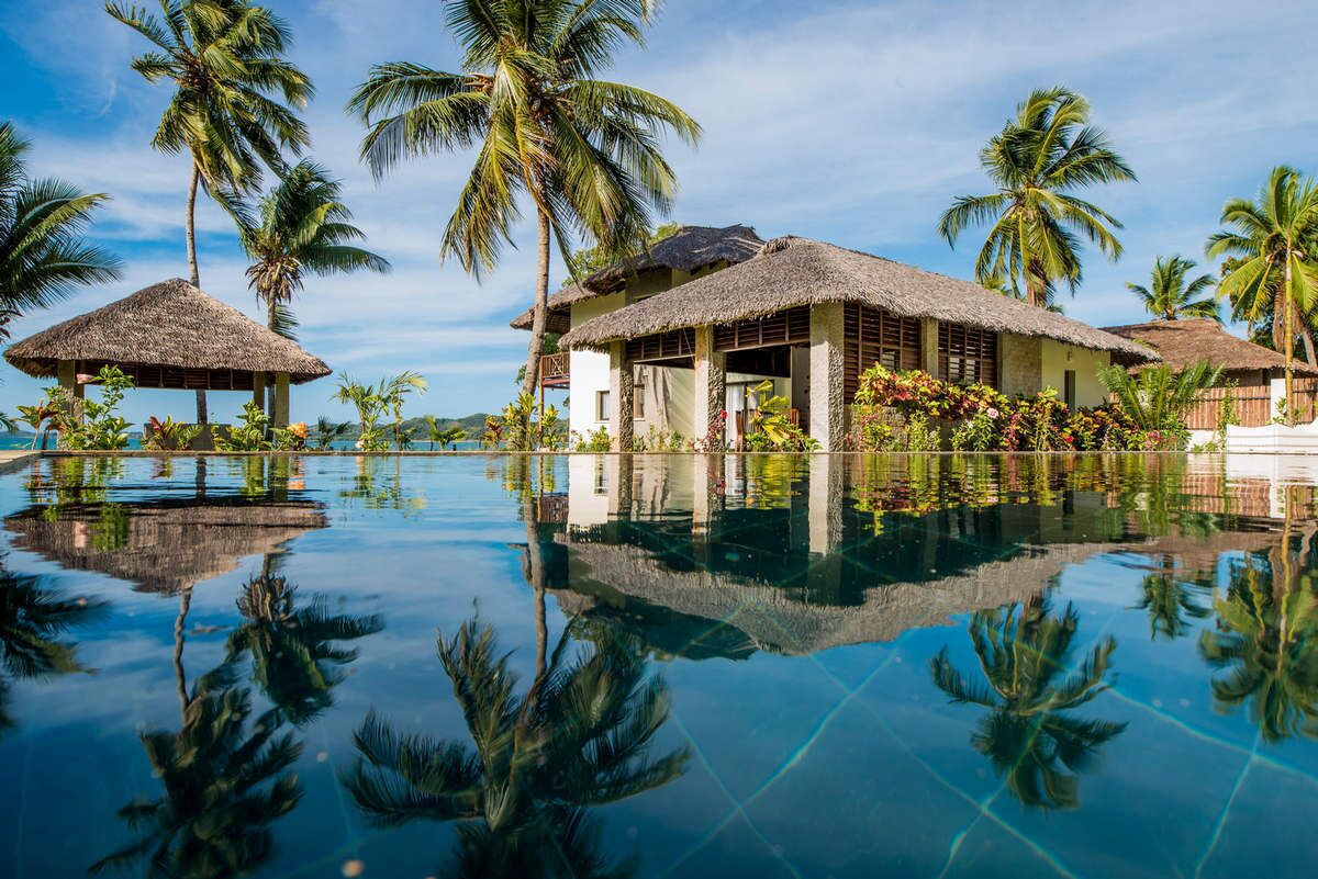 Un resort con edificios de techo de paja, piscina y palmeras.