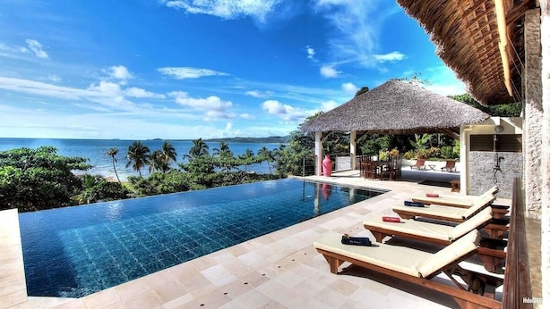 A infinity pool with a view of the ocean, lounge chairs, and a thatched-roof structure. - Home The Residence (Nosy Be)