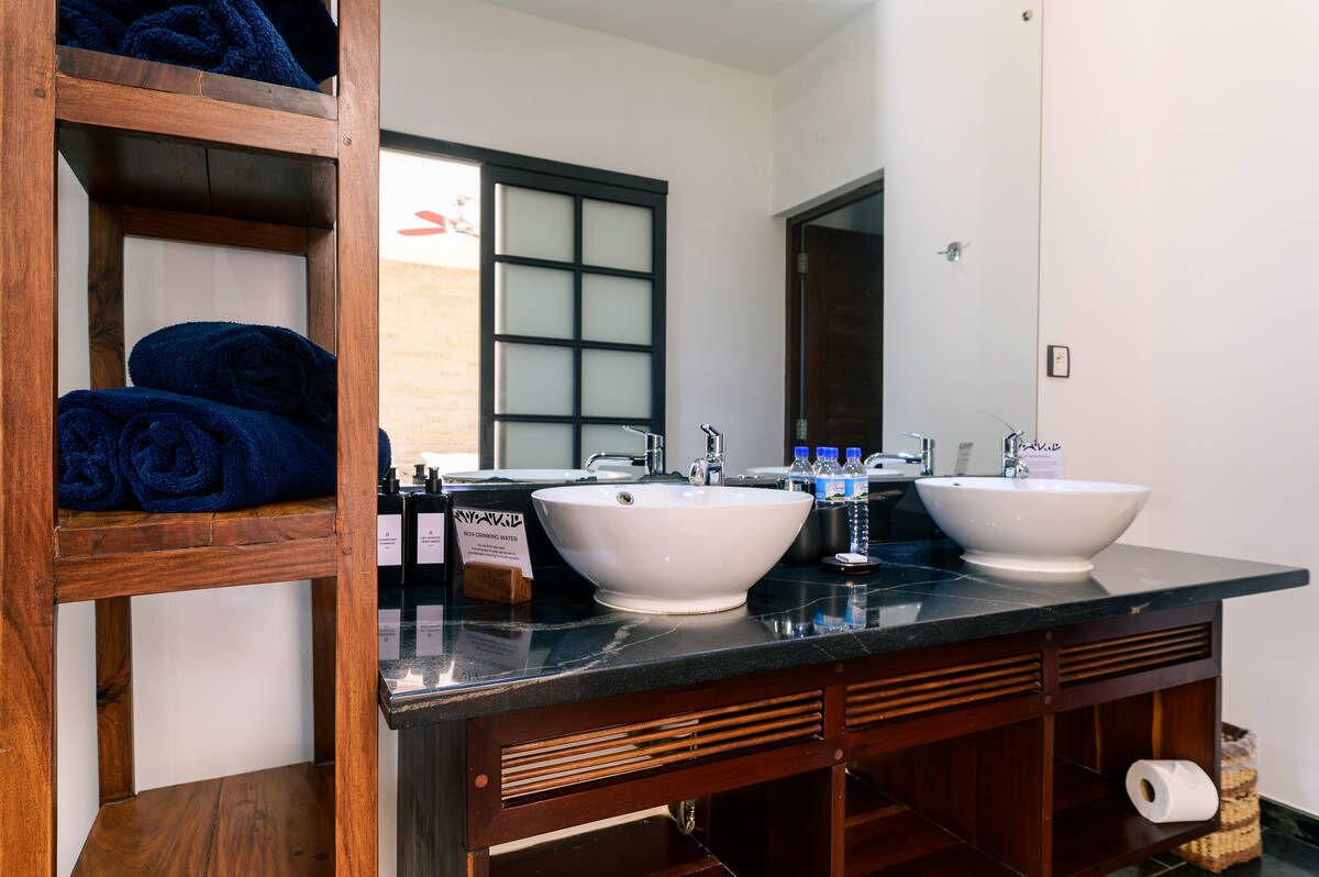 Un baño con dos lavamanos, un estante de madera con toallas y un espejo sobre los lavamanos.