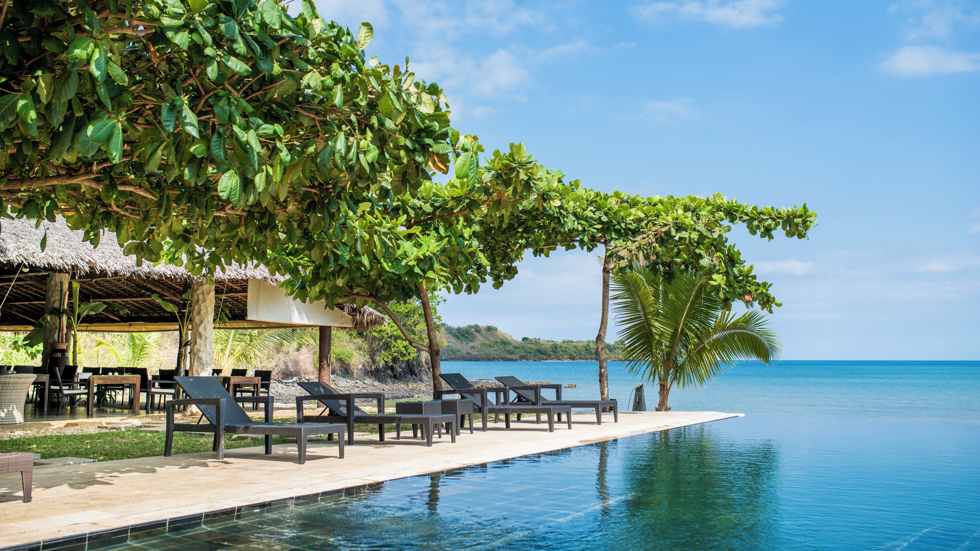 A poolside area with lounge chairs, a thatched-roof structure, and a view of the sea.