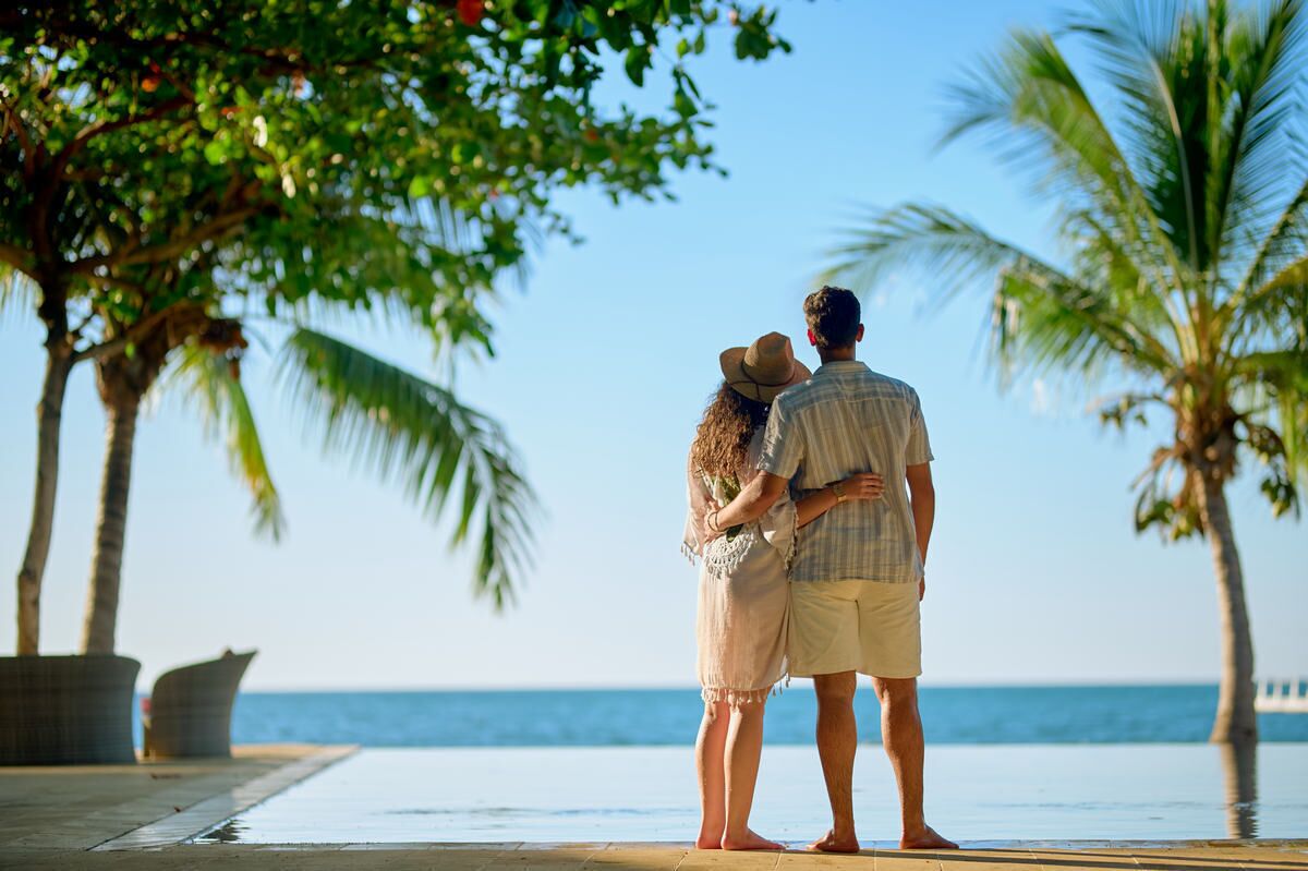A couple standing by a pool with palm trees and a view of the ocean.