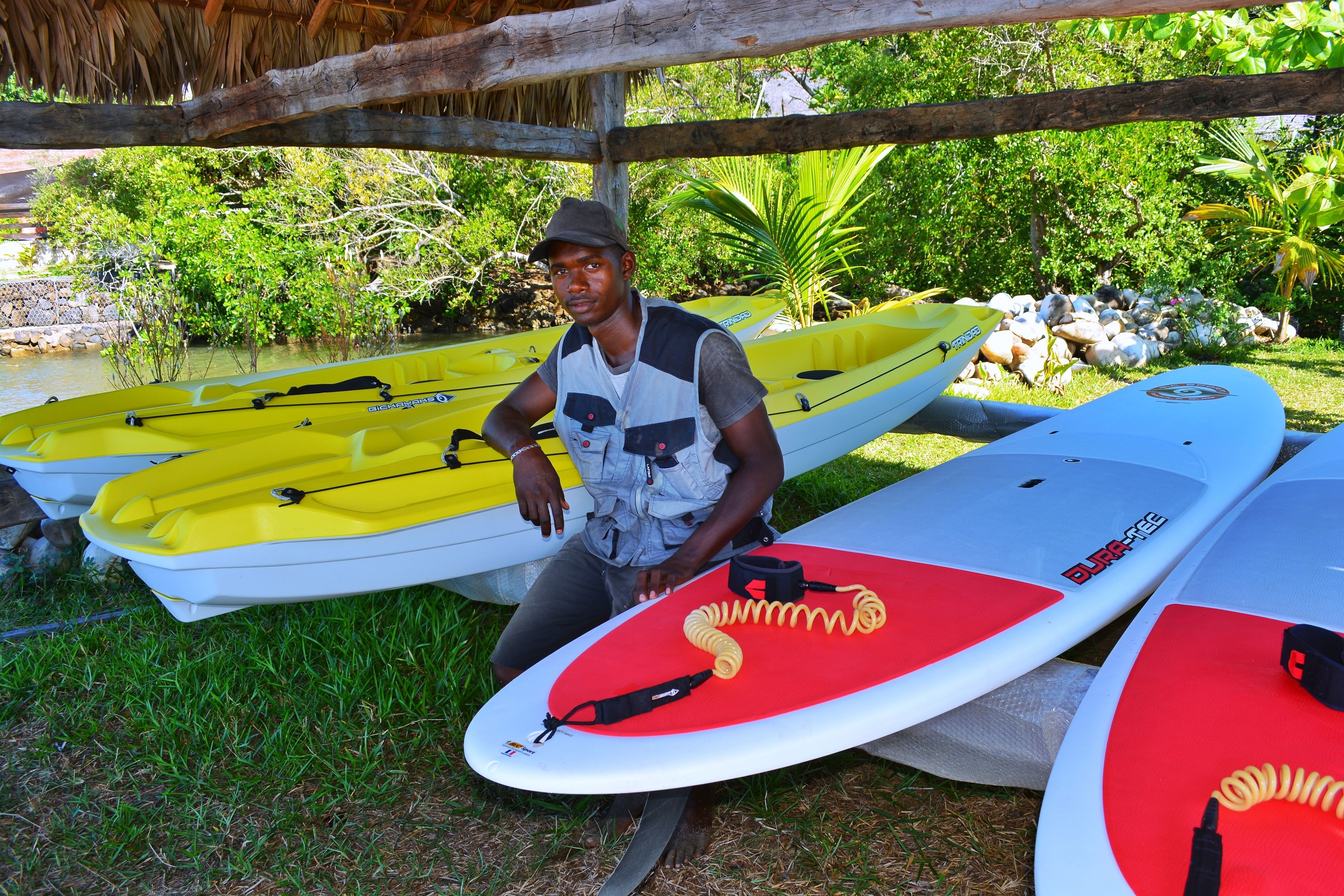 A man sitting next to a yellow kayak and a white surfboard with a red design.