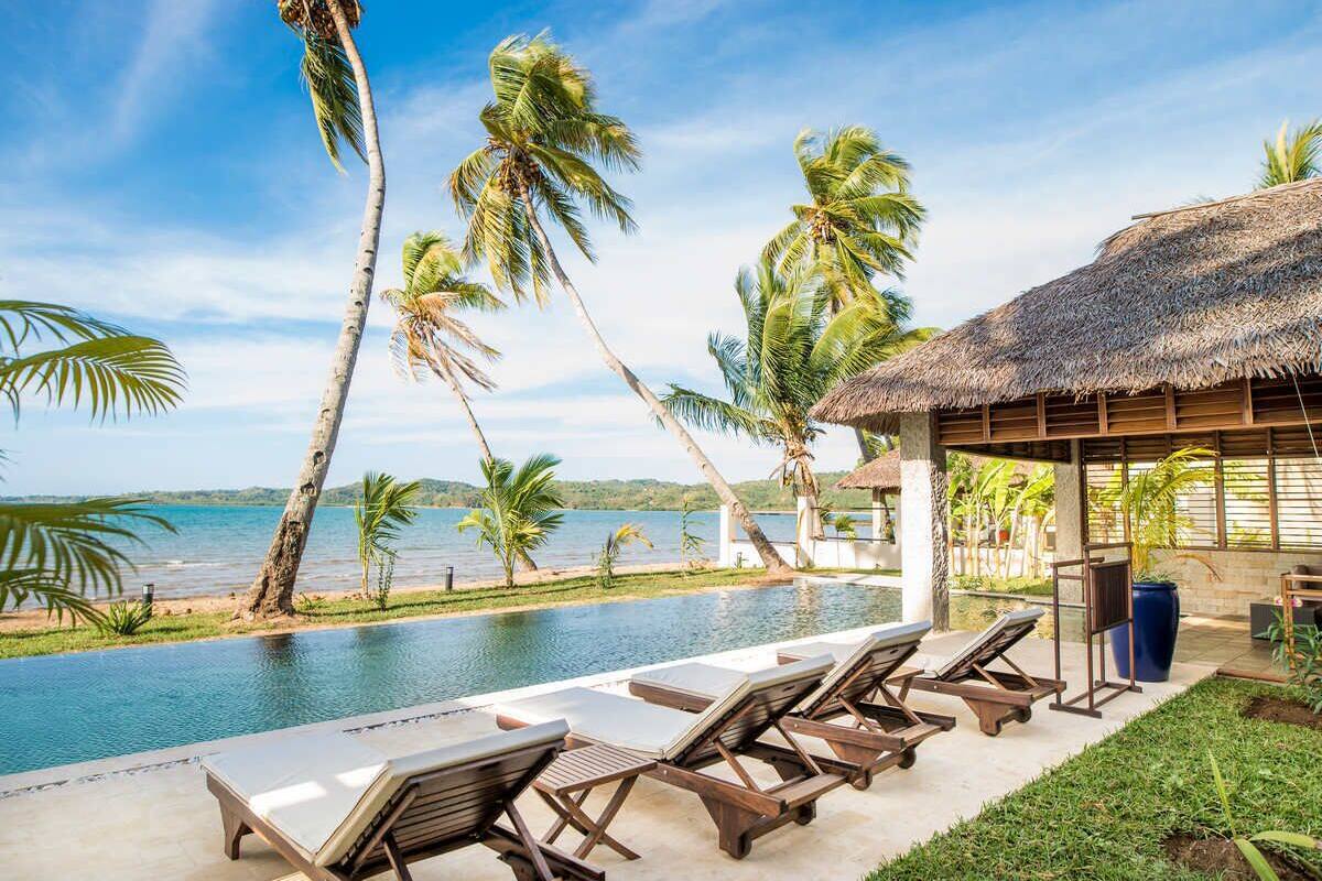 A poolside area with lounge chairs, a thatched-roof structure, and palm trees.
