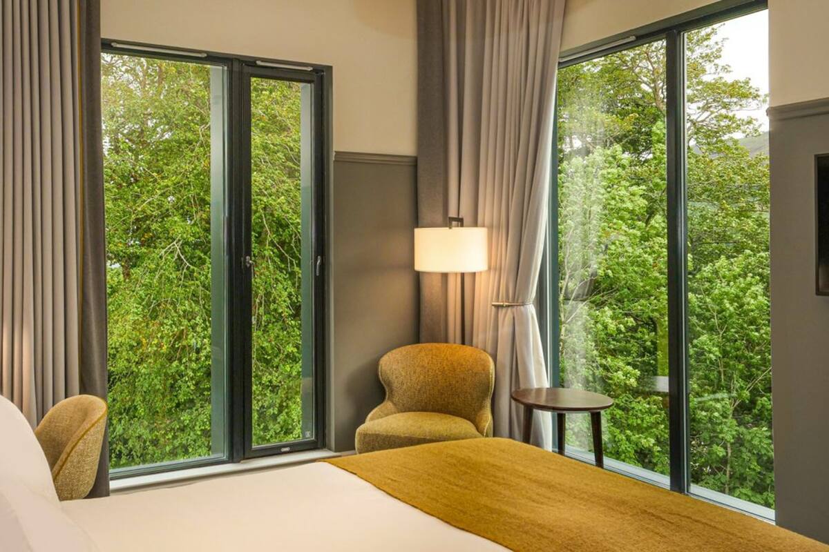 A hotel room with a bed, two chairs, a small table, and large windows offering a view of greenery.