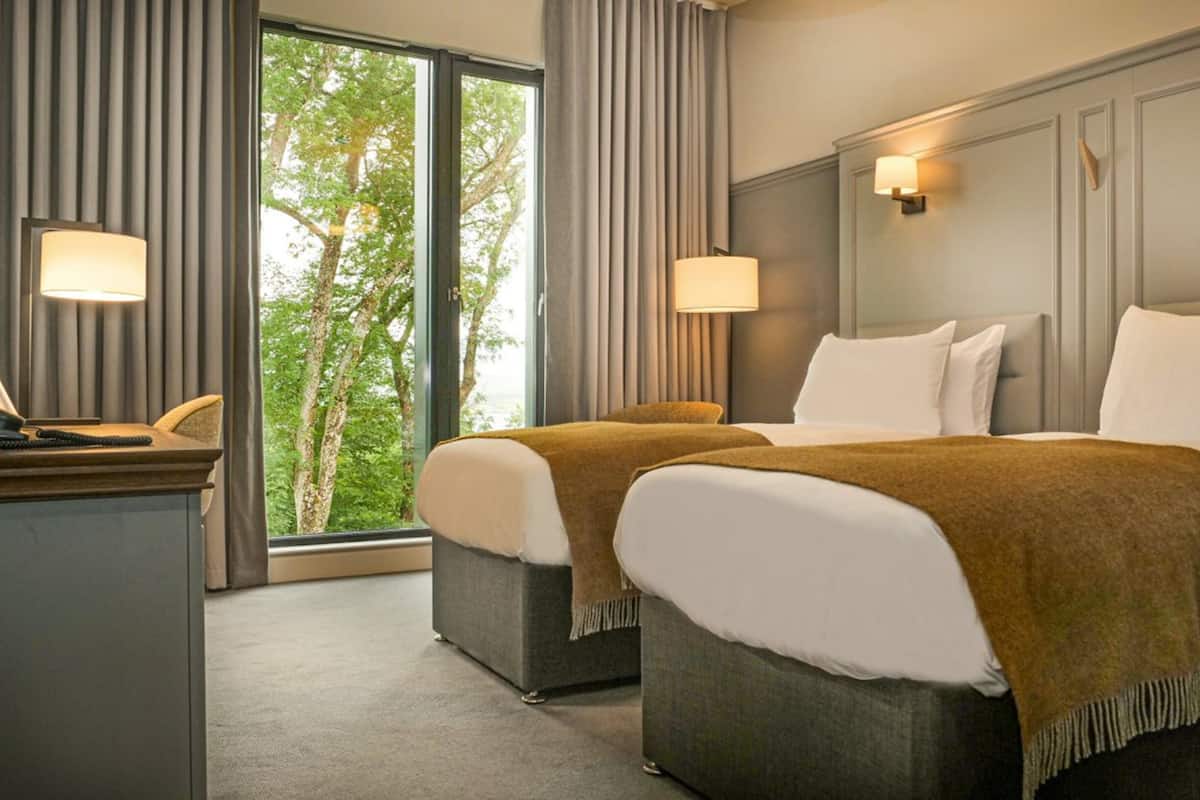 A hotel room with two beds, a desk, and a view of trees through large windows.