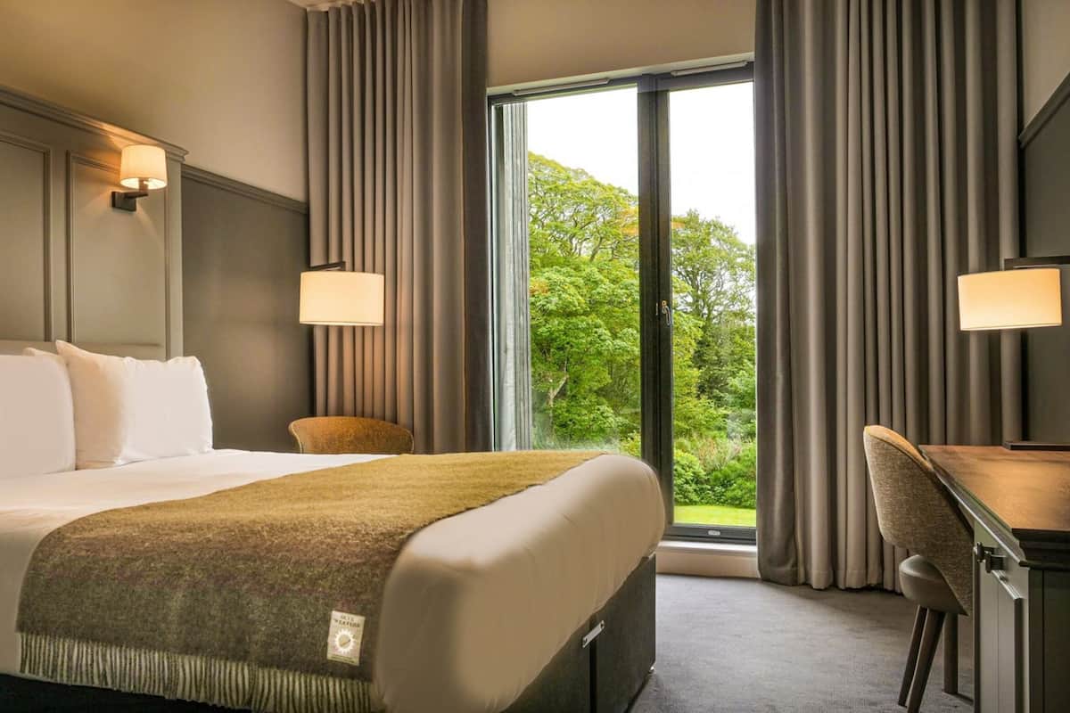 A hotel room with a large bed, two bedside lamps, a desk, and a view of greenery through the window.
