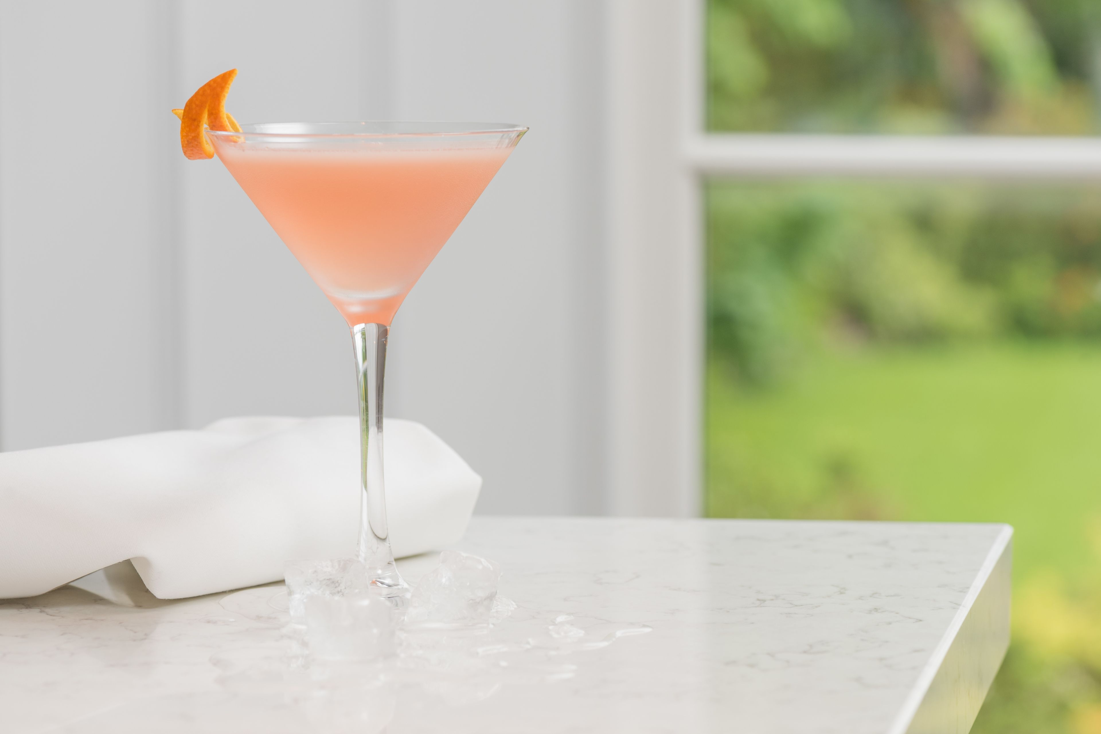 A pink cocktail in a martini glass with an orange garnish, placed on a marble surface with a white napkin beside it.