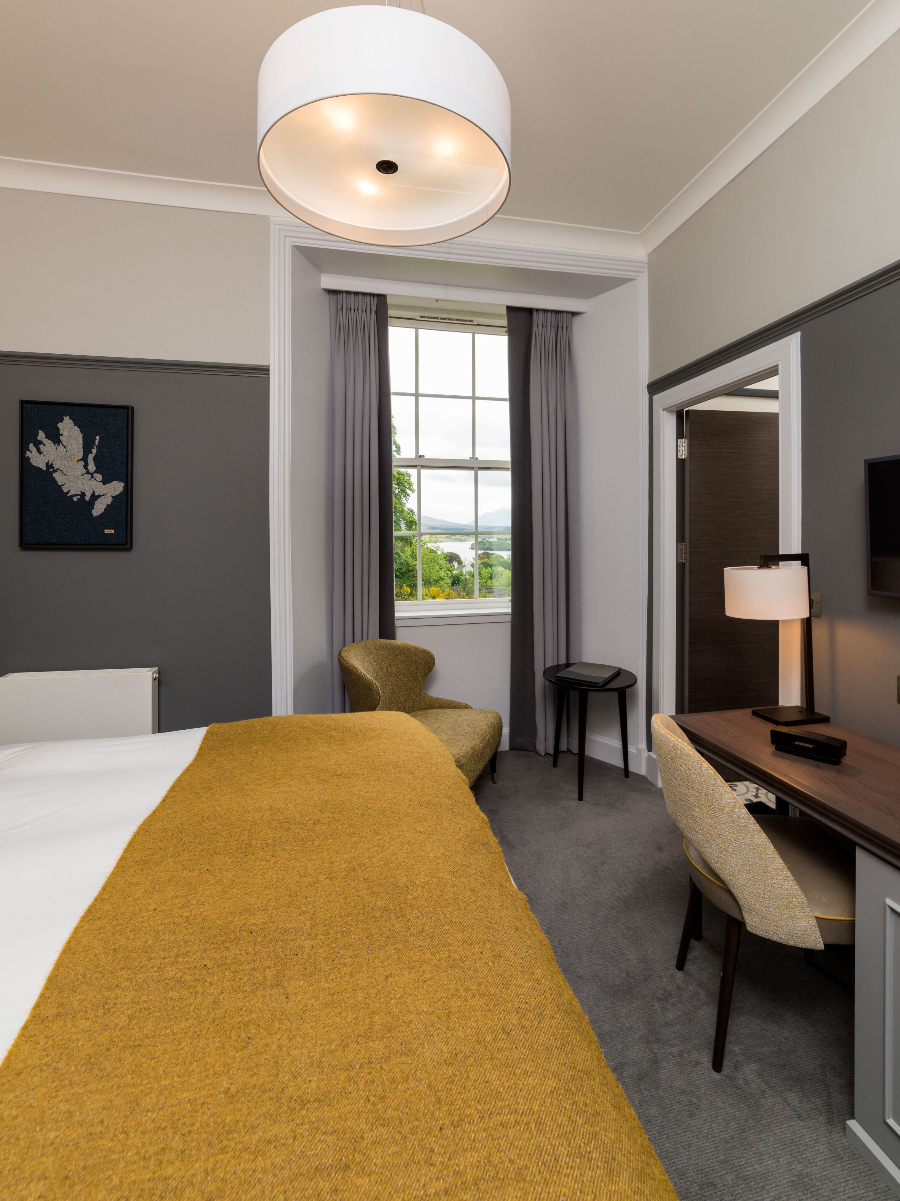 Una habitación de hotel con una cama grande, un escritorio, una silla, una lámpara, un ventanal con cortinas y un mapa enmarcado en la pared.