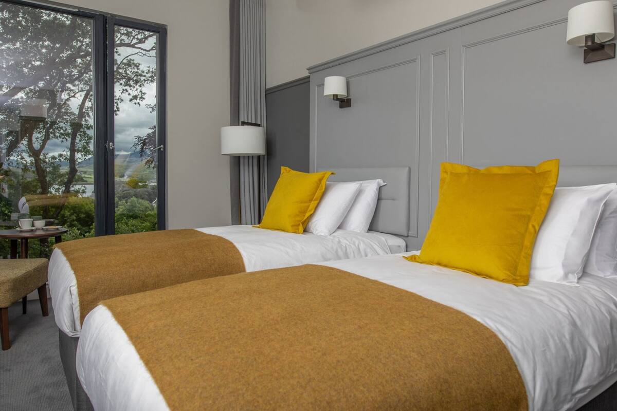 A hotel room with two beds, each with white bedding and a mustard-colored throw blanket. There are two bedside lamps, a window with a view of trees and hills, and a small table with a chair.